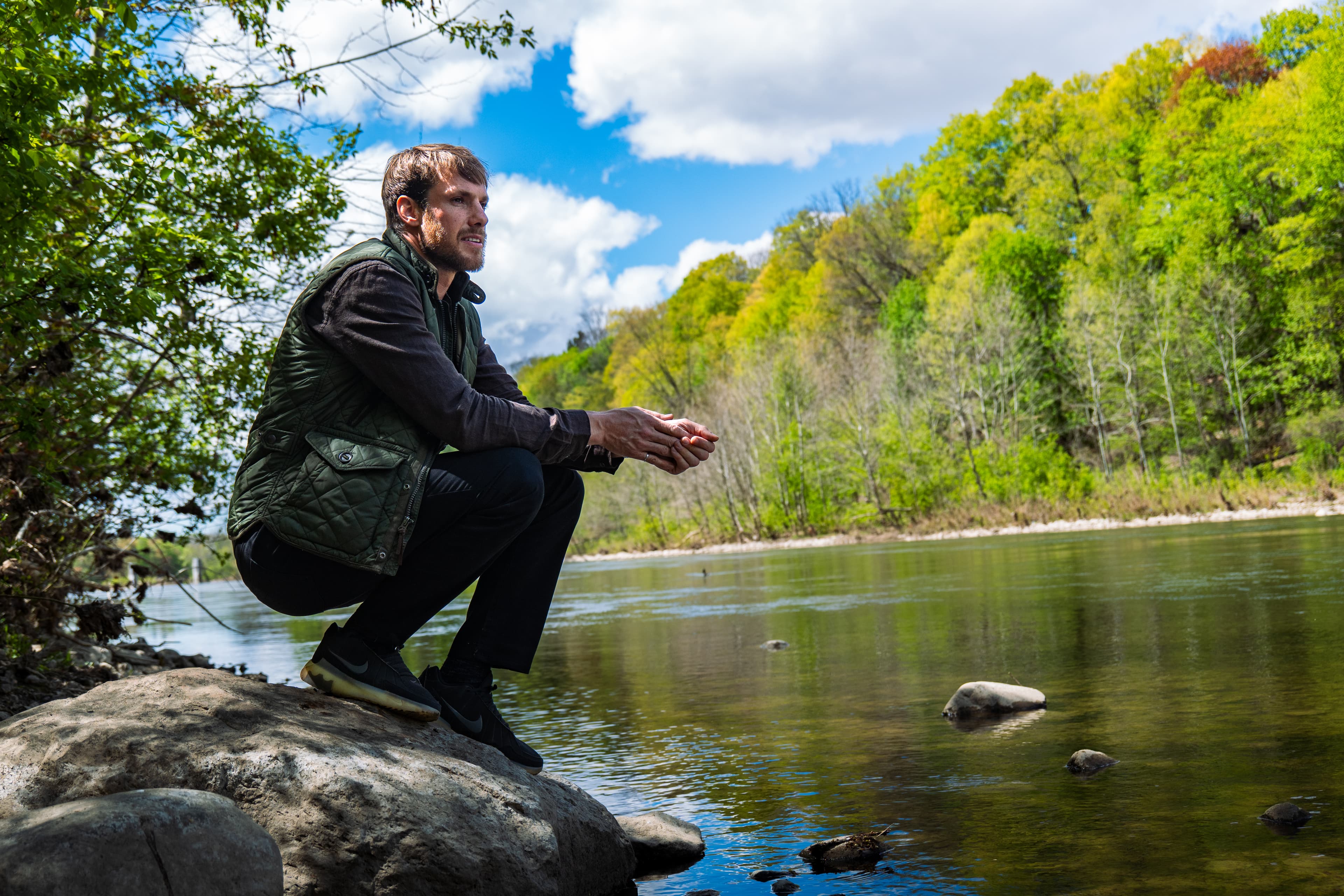 Man crouched on a rock beside a river under blue sky