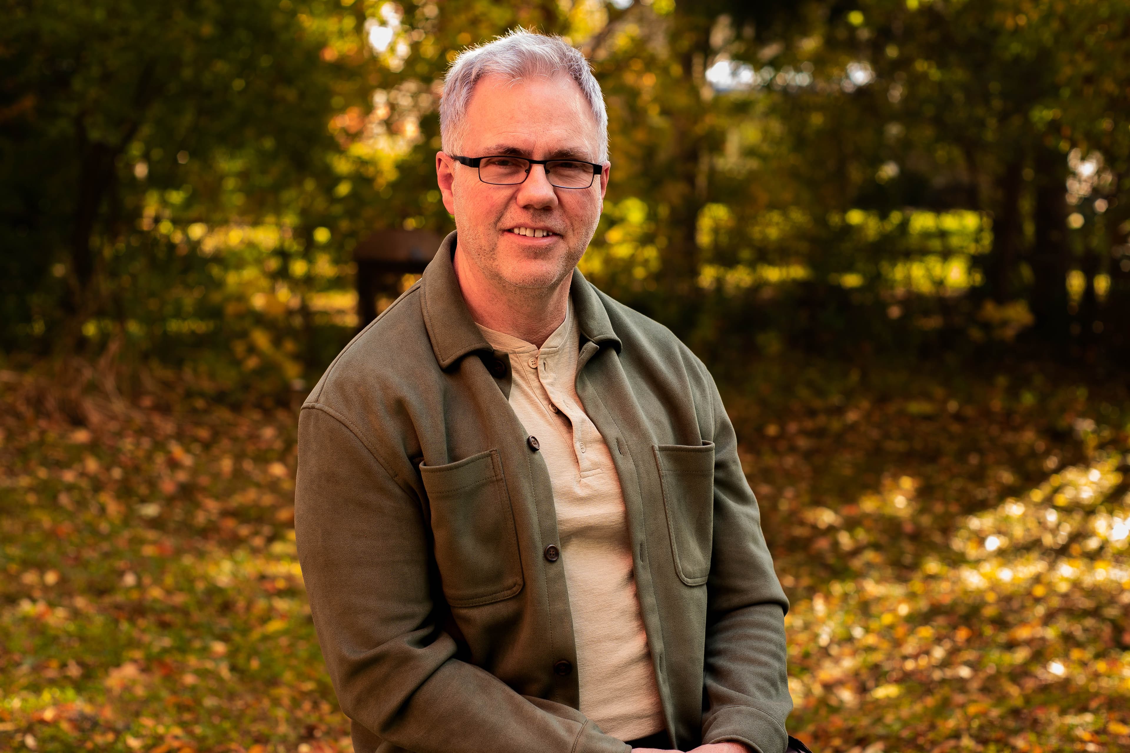 An older gentleman in a green jacket smiling in an autumn forest